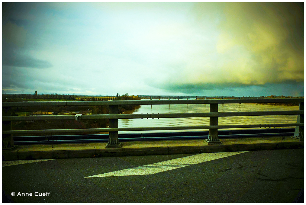 Depuis le pont, le regard glisse entre Garonne et Dordogne. À gauche, l’eau pulse en reflets d’étain ; à droite, elle s’étire en velours sombre. Le ciel, ni bleu ni gris, semble peint à la tempera, chaque nuage posé comme une touche hésitante. Les berges s’effacent dans une brume dorée, et les arbres deviennent esquisses. Ce n’est plus une photographie. C’est une toile qui respire. Un instant suspendu entre deux Rives.